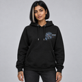 A woman modelling a Deshino black pullover hoodie with a detailed 'After Midnight' graphic print on the left chest, paired with dark grey jeans, against a solid grey studio background.