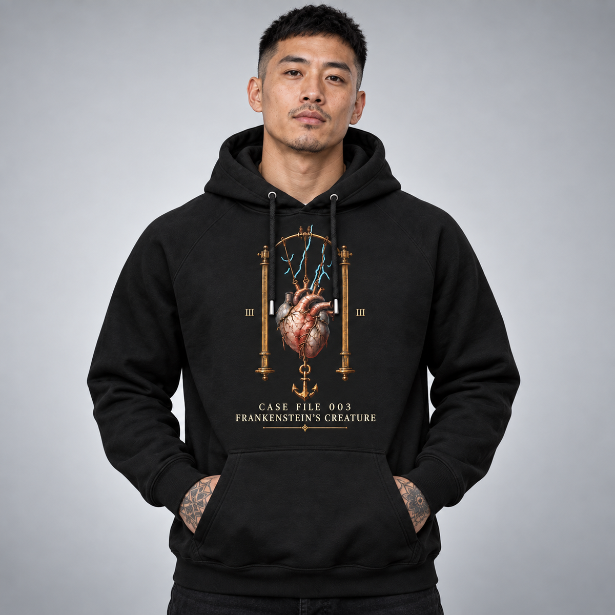 A man wearing a black pullover hoodie with a detailed graphic print of an anatomical heart, an anchor, and gold Roman numerals and text reading 'CASE FILE 003 FRANKENSTEIN'S CREATURE' on the front, set against a plain grey studio background.
