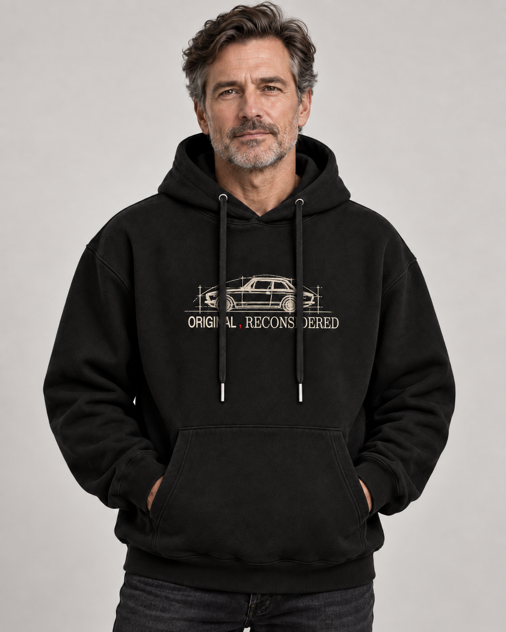 A man wearing a black pullover hoodie with a beige graphic of a station wagon and the text 'ORIGINAL RECONSIDERED' on the chest, posed against a neutral gray studio background.