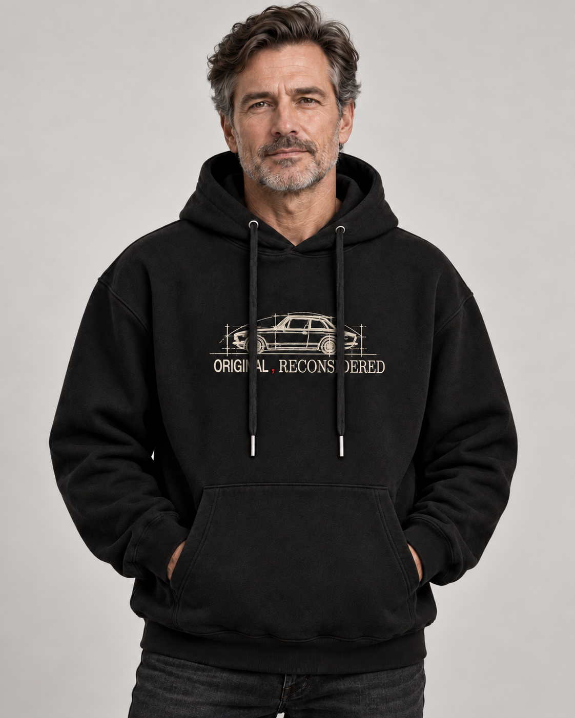 A man wearing a black pullover hoodie with a beige graphic of a station wagon and the text 'ORIGINAL RECONSIDERED' on the chest, posed against a neutral gray studio background.