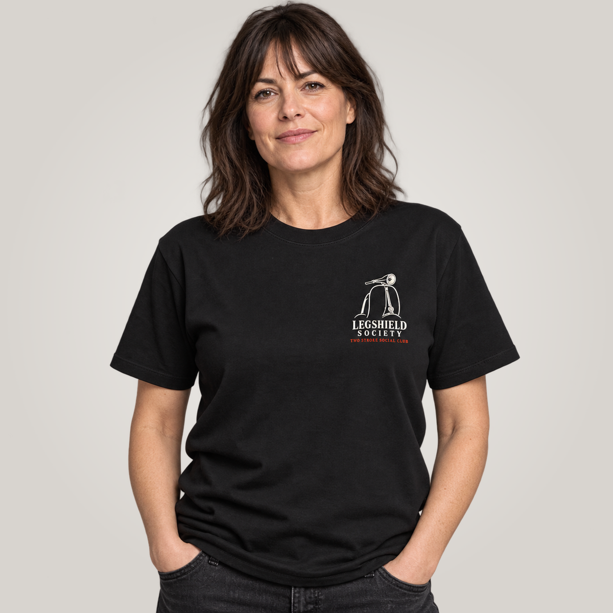A woman wearing a black crewneck t-shirt with a small white and red 'Legshield Society' logo on the left chest, photographed against a solid light grey background.