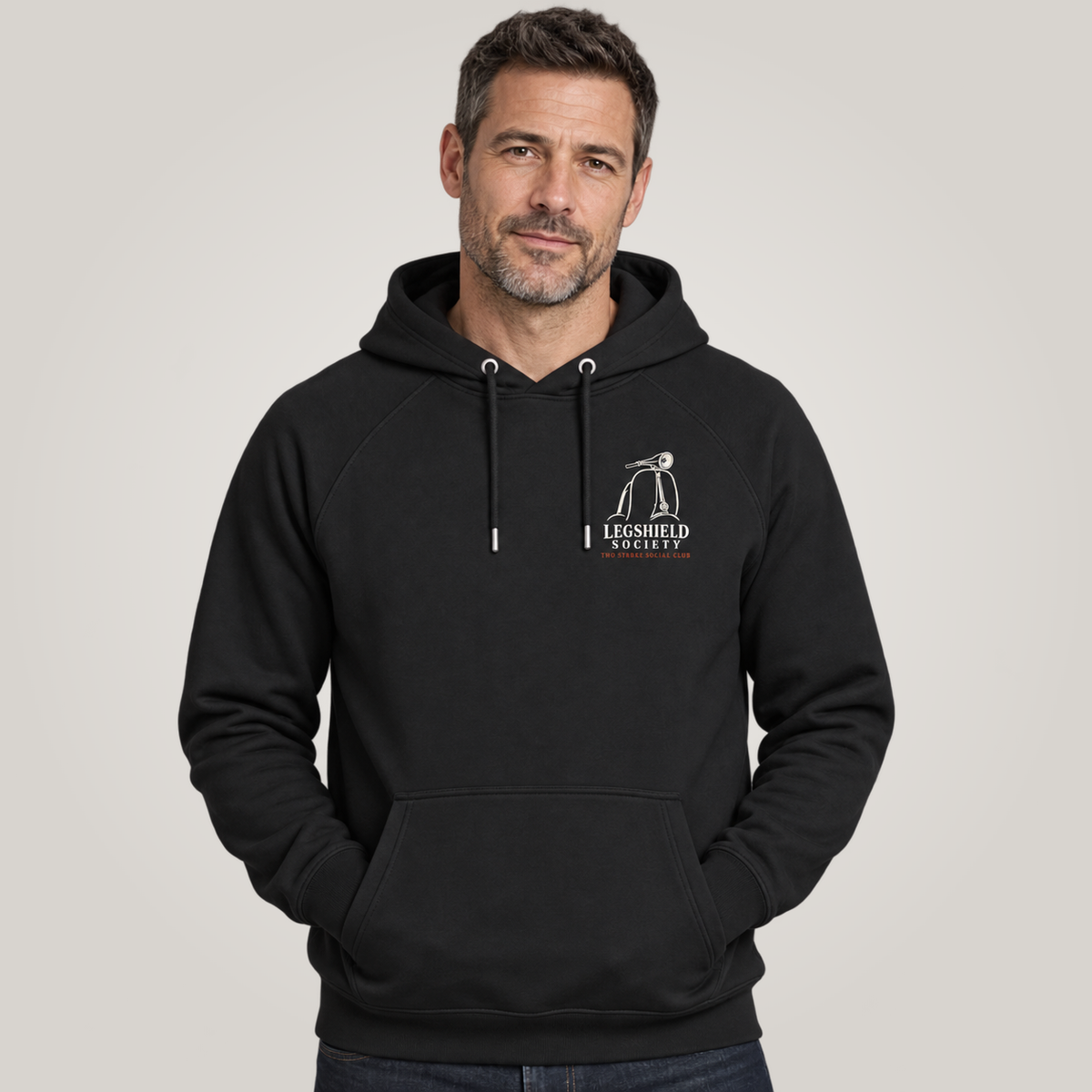 A man wearing a black pullover hoodie with a white 'Legshield Society' logo on the left chest, set against a plain light gray studio background.