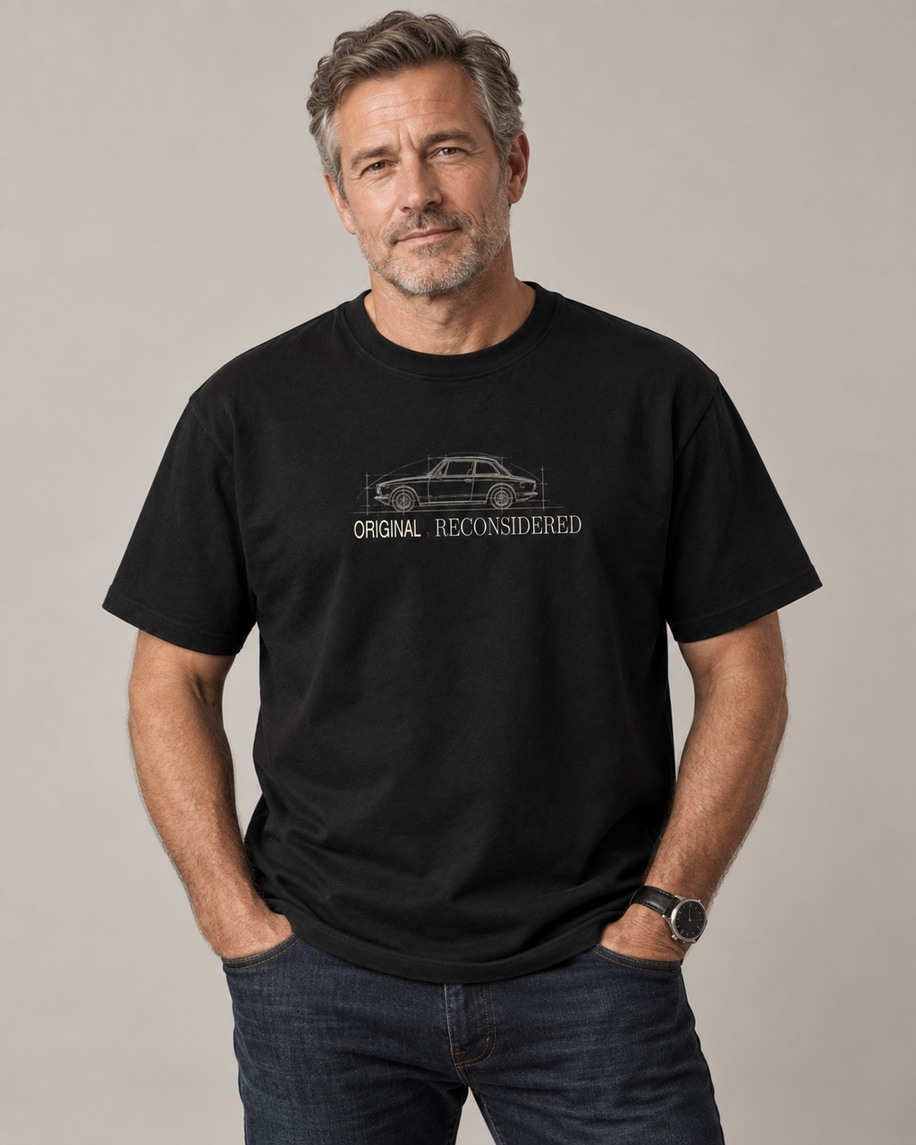 A man wearing a black short-sleeve t-shirt with a white line art graphic of a classic car and the text 'ORIGINAL RECONSIDERED' on the chest, paired with dark blue jeans, against a neutral gray background.