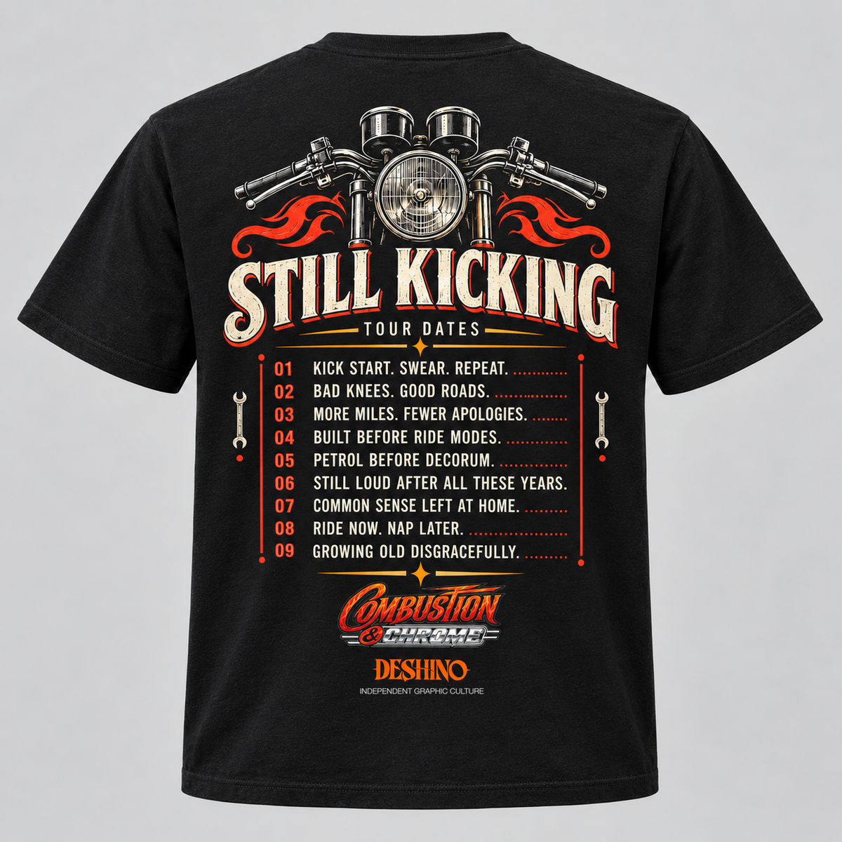 A black short-sleeve t-shirt with a large back graphic featuring motorcycle handlebars, red flames, and the text 'Still Kicking Tour Dates' above a numbered list of locations and a 'Combustion & Chrome' & 'Deshino' logo, shown on a light gray background.