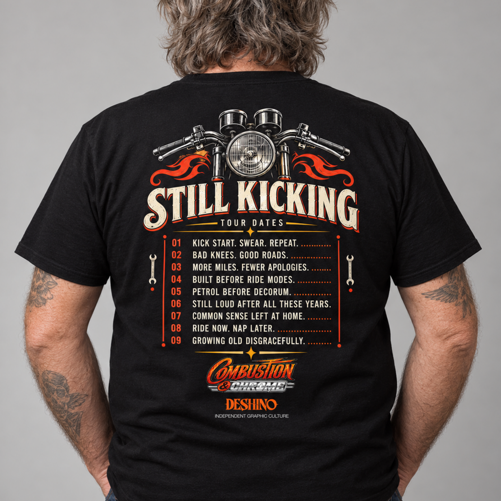 A man wearing a black t-shirt, viewed from the back. The shirt features a large graphic print with a motorcycle engine, flames, the text 'STILL KICKING TOUR DATES', a numbered list, and the 'Combustion & Chrome' & 'Deshino' logos.