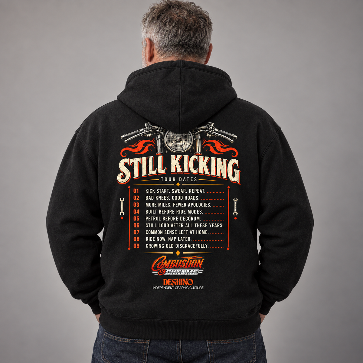 A man viewed from behind wearing a black hoodie with a large back graphic. The graphic features a motorcycle illustration, the text 'STILL KICKING TOUR DATES', a numbered list, and the 'Combustion & Chrome' and 'Deshino' logos.