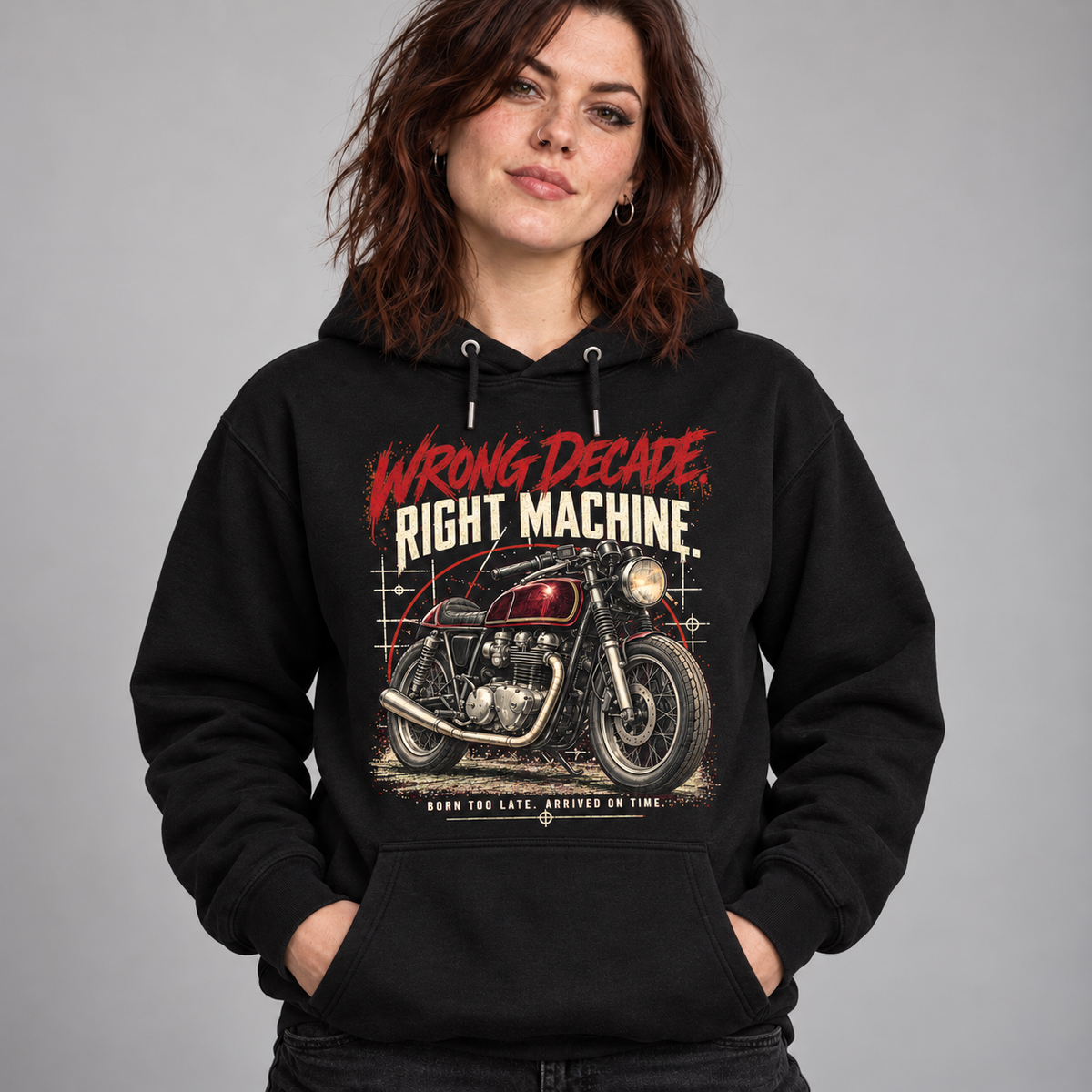 A woman wearing a black pullover hoodie with a vintage-style graphic of a red motorcycle and the text 'WRONG DECADE, RIGHT MACHINE' on a plain gray studio background.